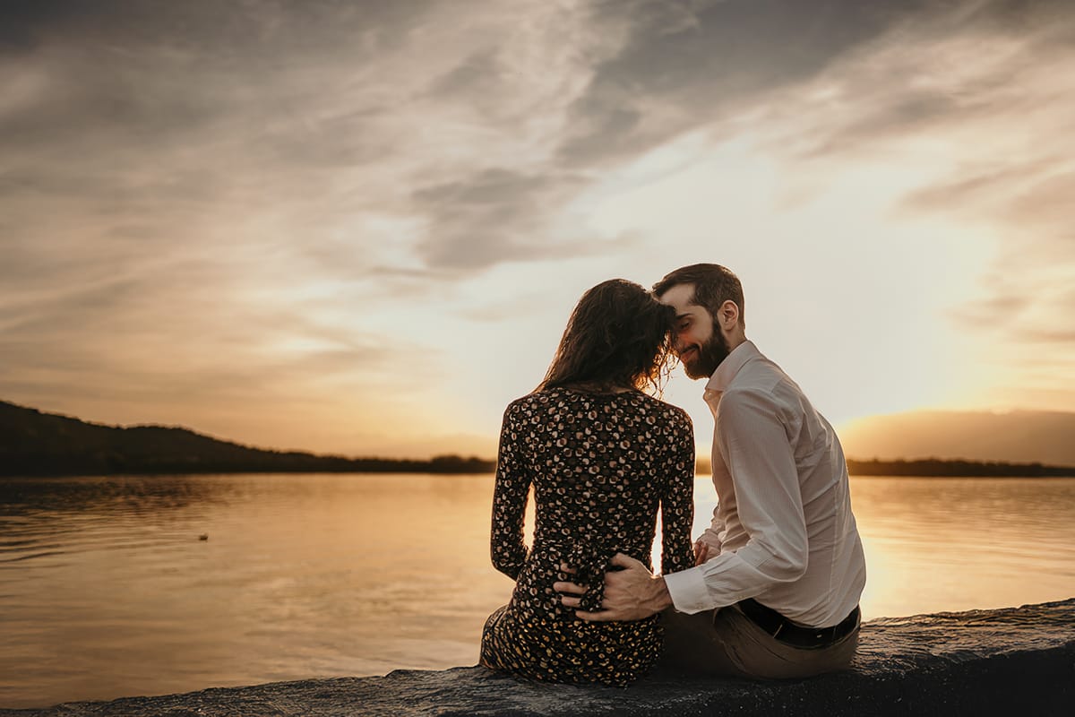 Pre-wedding al Lago di Viverone di Ilaria e Luca. La coppia si guarda teneramente mentre è seduta sul pontile in pietra davanti al bellissimo tramonto sul lago.