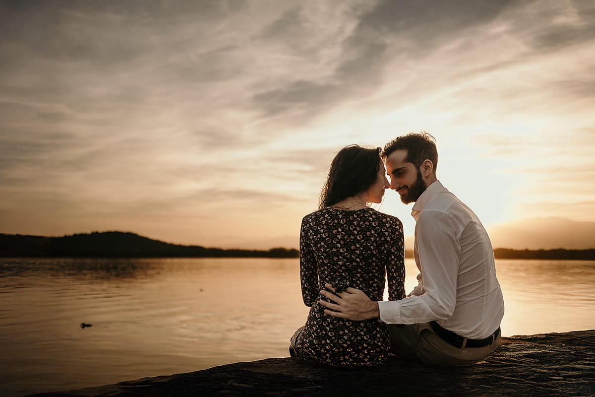 Pre-wedding al Lago di Viverone di Ilaria e Luca. La coppia si guarda teneramente mentre è seduta sul pontile in pietra davanti al bellissimo tramonto sul lago.