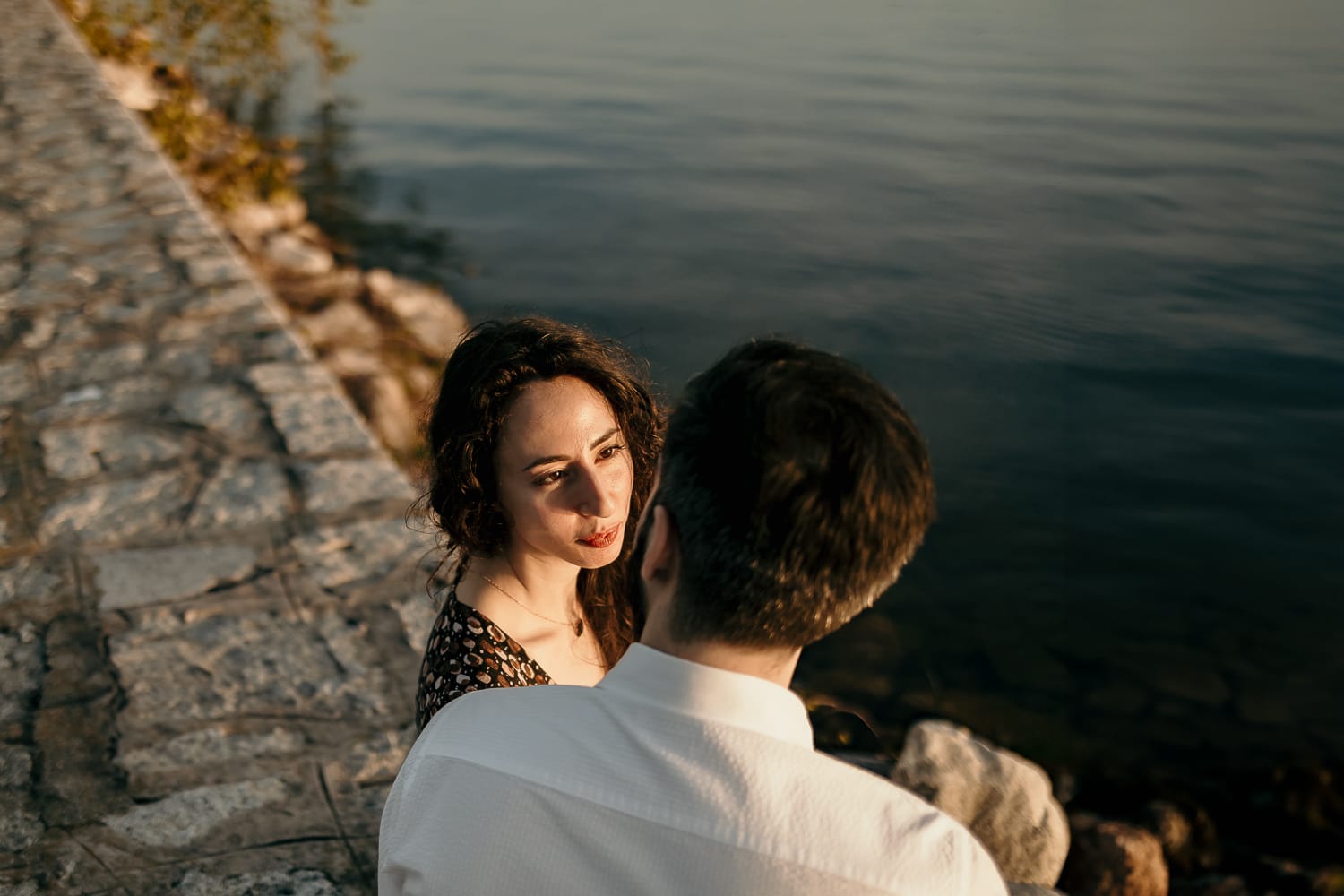 Pre-wedding al Lago di Viverone di Ilaria e Luca. La coppia si guarda teneramente mentre è seduta sul pontile in pietra davanti al bellissimo tramonto sul lago.