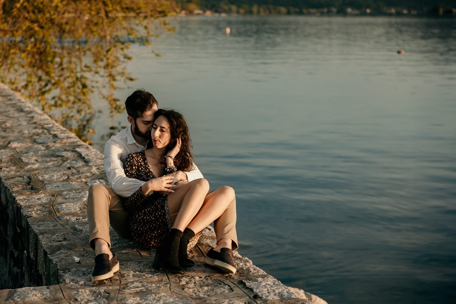 Pre-wedding al Lago di Viverone di Ilaria e Luca. la coppia è seduta sul pontile di pietra e Ilaria è tra le braccia di Luca.