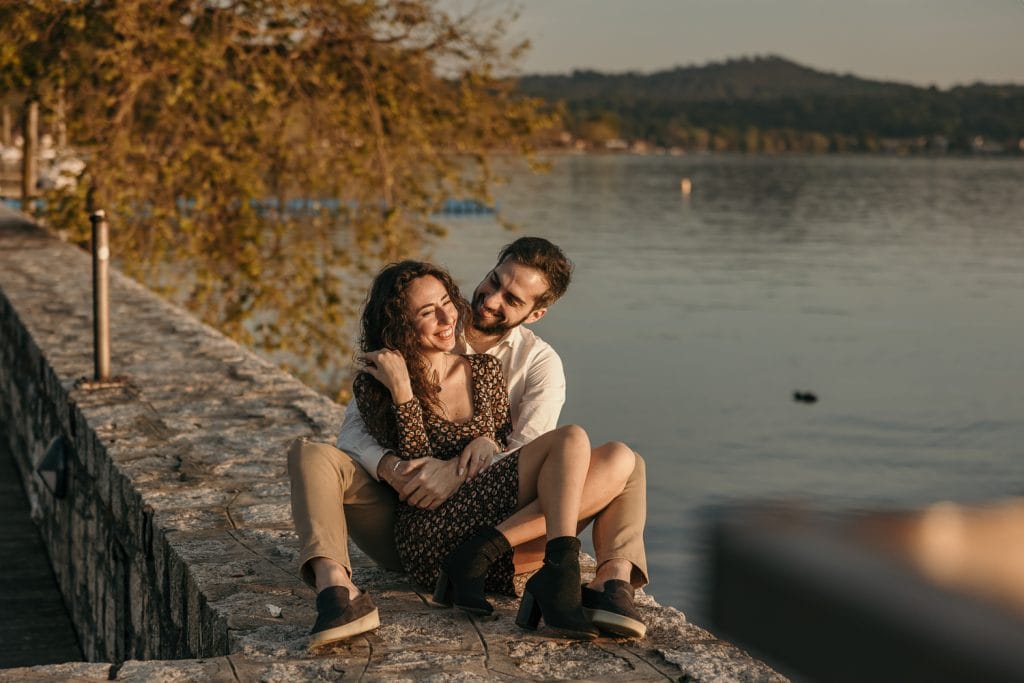 Ilaria and Luca's pre-wedding shoot at Lake Viverone. The couple is sitting on a stone pier, and Ilaria is in Luca's arms. They are smiling.  