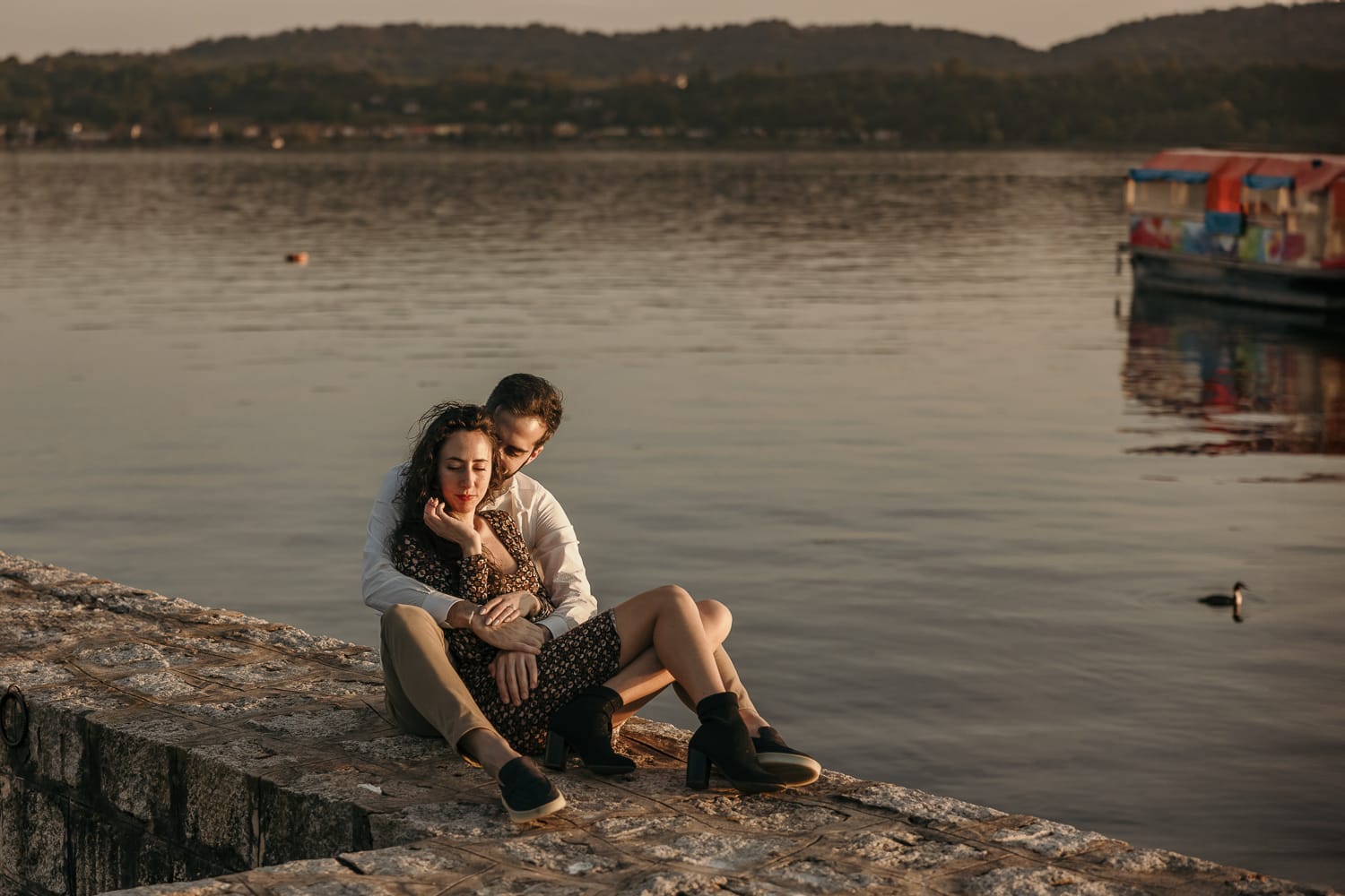 Pre-wedding al Lago di Viverone di Ilaria e Luca. la coppia è seduta sul pontile di pietra e Ilaria è tra le braccia di Luca. Si guardano