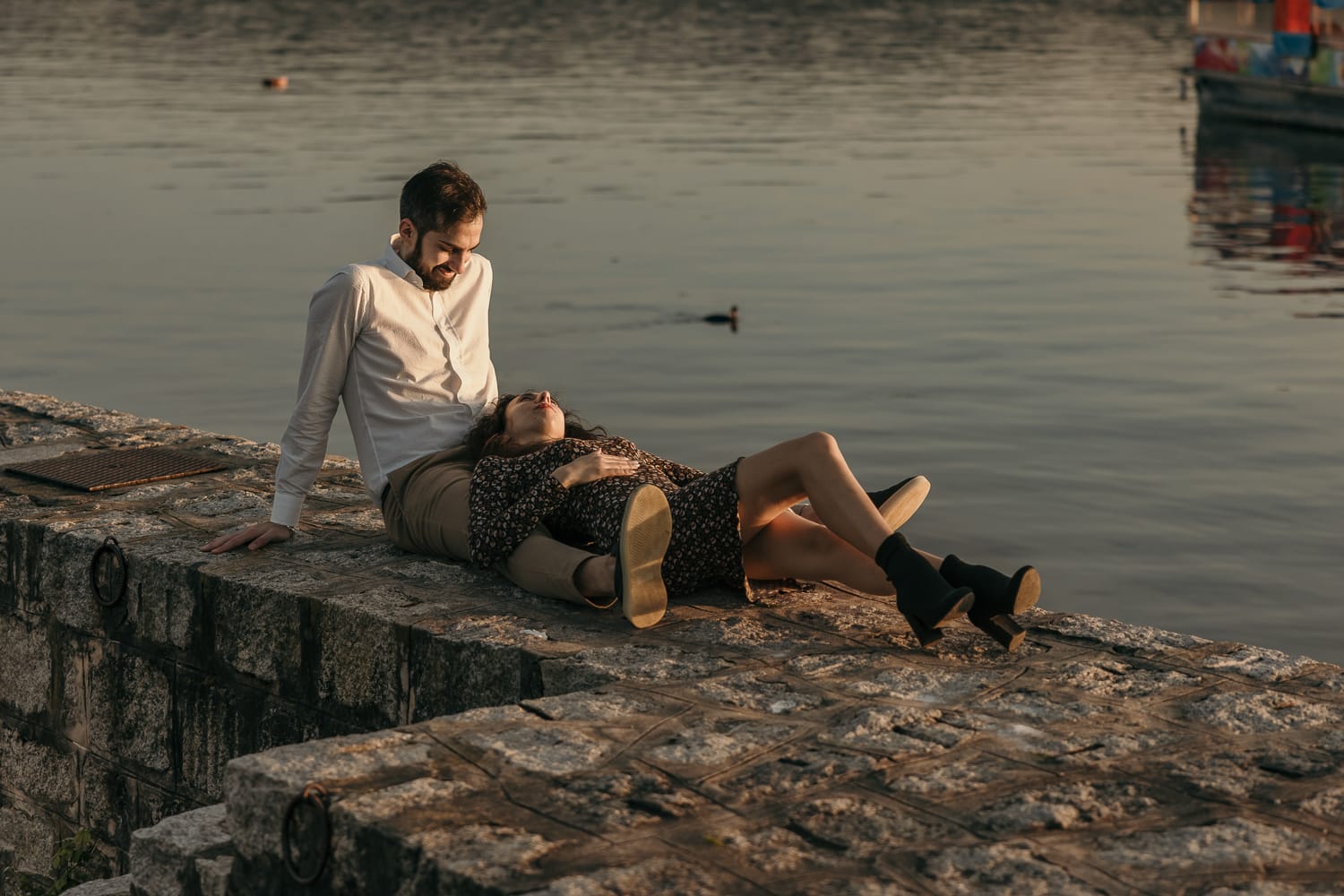 Pre-wedding al Lago di Viverone di Ilaria e Luca. la coppia è seduta sul pontile di pietra e Ilaria è tra le braccia di Luca. Ilaria è sdraiata.