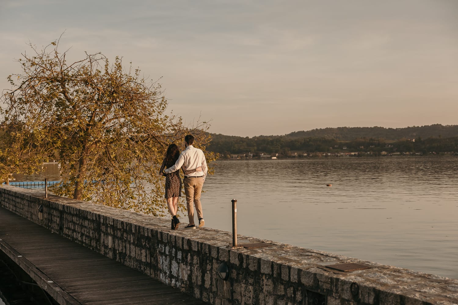 Pre-wedding al Lago di Viverone di Ilaria e Luca. La coppia cammina abbracciata sul pontile di pietra sul lago.