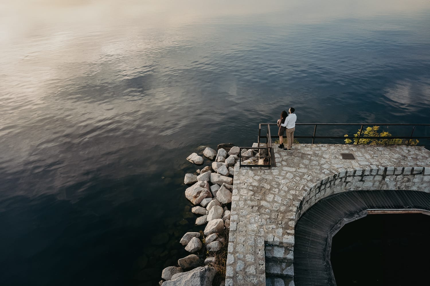 Pre-wedding al Lago di Viverone di Ilaria e Luca. Foto aerea dal drone della coppia sul pontile di pietra sul lago che guarda il lago.