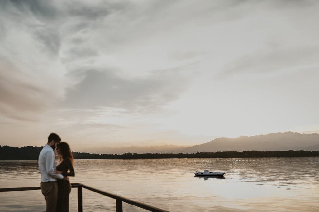 Ilaria and Luca's love story unfolds against the picturesque backdrop of Lake Viverone. In this pre-wedding photo, the couple stands in the foreground, their eyes locked on each other, as a boat glides peacefully across the water. 