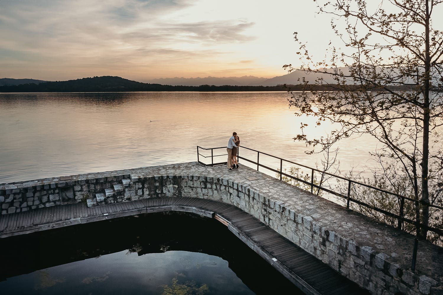Pre-wedding al Lago di Viverone di Ilaria e Luca. Foto aerea dal drone della coppia sul pontile di pietra. Si vede bene il bellissimo tramonto sul lago.