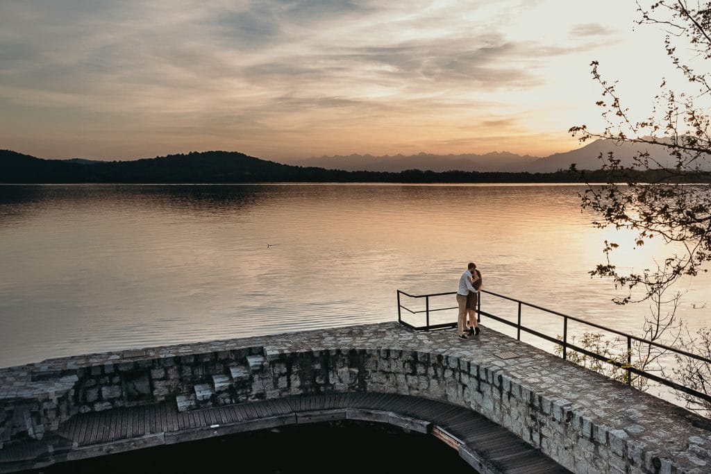 Ilaria and Luca's love story is captured in this aerial drone photo taken at Lake Viverone during their pre-wedding shoot. The couple, standing on the stone pier, is bathed in the warm glow of the setting sun.  
