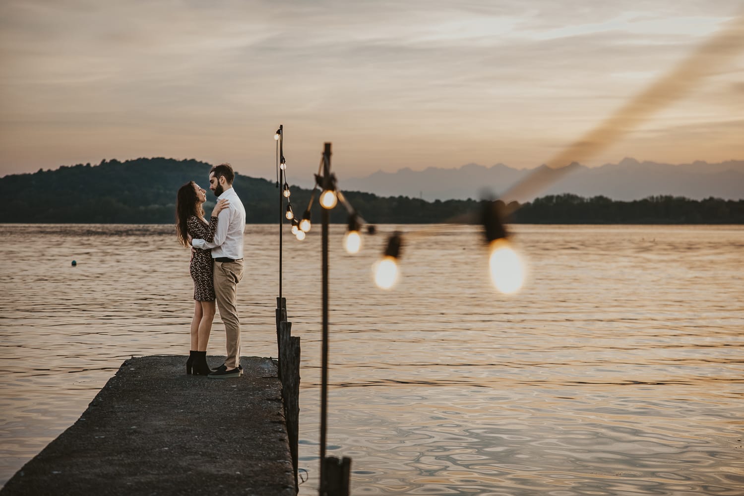 Pre-wedding al Lago di Viverone di Ilaria e Luca. Sposi abbracciati sul pontile di legno decorato da una fila di lampadine. Sullo sfondo un bellissimo tramonto.