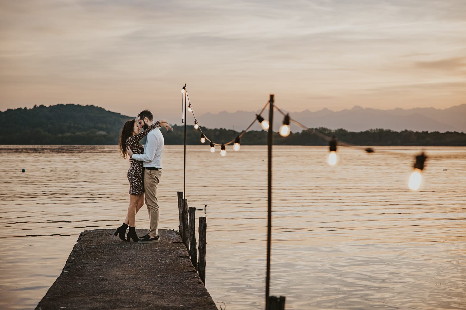 Pre-wedding al Lago di Viverone di Ilaria e Luca. Sposi abbracciati sul pontile di legno decorato da una fila di lampadine. Sullo sfondo un bellissimo tramonto.