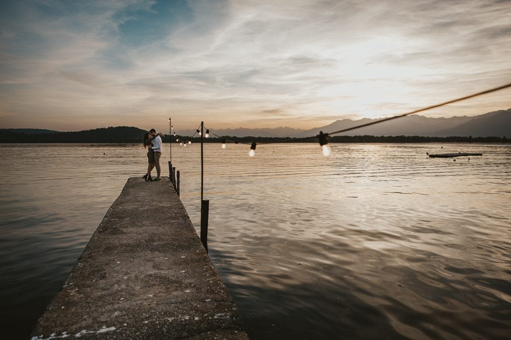 Ilaria and Luca's pre-wedding shoot at Lake Viverone. The couple is hugging on a wooden pier decorated with a row of light bulbs. A beautiful sunset is in the background.  