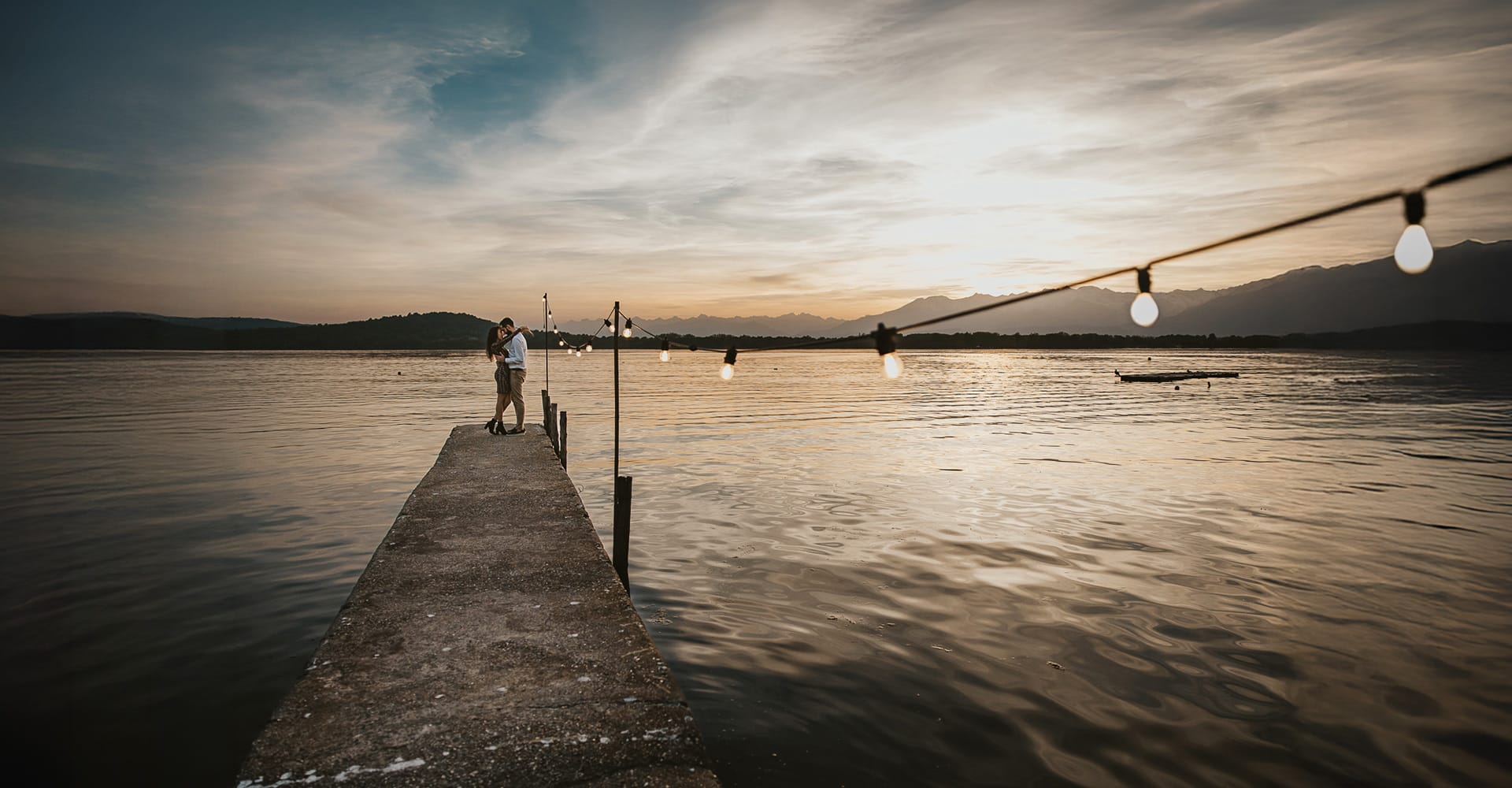 Pre-wedding al Lago di Viverone di Ilaria e Luca. Sposi sul molo di legno con un fila di luci accese e uno splendido tramonto sul lago sullo sfondo.