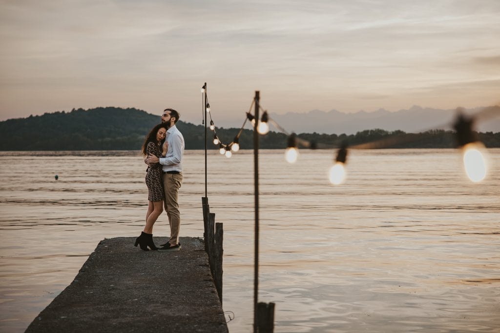 Ilaria and Luca's pre-wedding shoot at Lake Viverone. The couple is hugging on a wooden pier decorated with a row of light bulbs. A beautiful sunset is in the background.  
