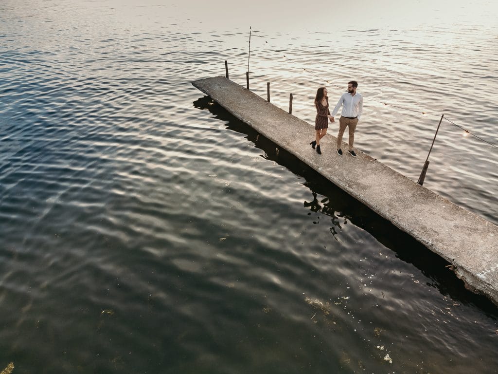 Ilaria and Luca's pre-wedding shoot at Lake Viverone. An aerial drone shot captures the couple embracing on a wooden pier adorned with fairy lights against a stunning sunset.   