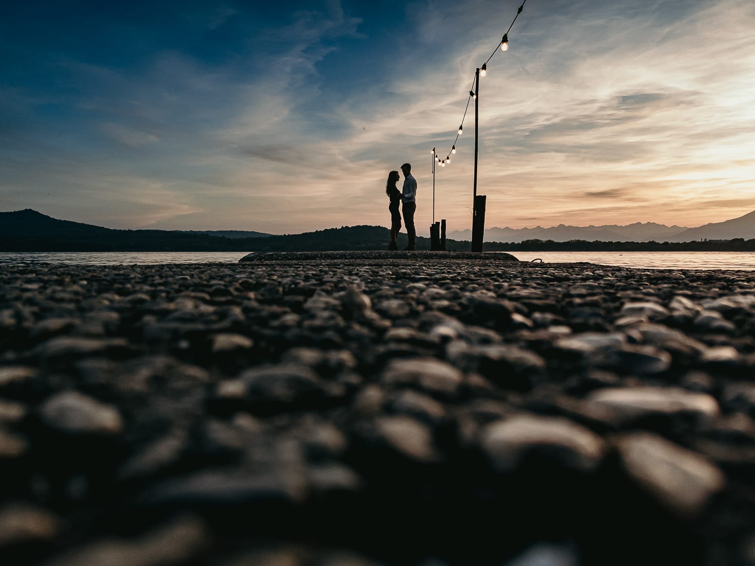 Pre-wedding al Lago di Viverone di Ilaria e Luca. Sposi abbracciati sul pontile di legno decorato da una fila di lampadine. Sullo sfondo un bellissimo tramonto. Foto da drone messo per terrra.