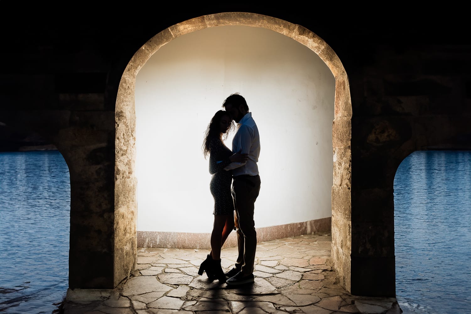 Pre-wedding al Lago di Viverone di Ilaria e Luca. Silhouette della coppia sotto un portico sul lago. Uso del flash off camera dietro di loro.