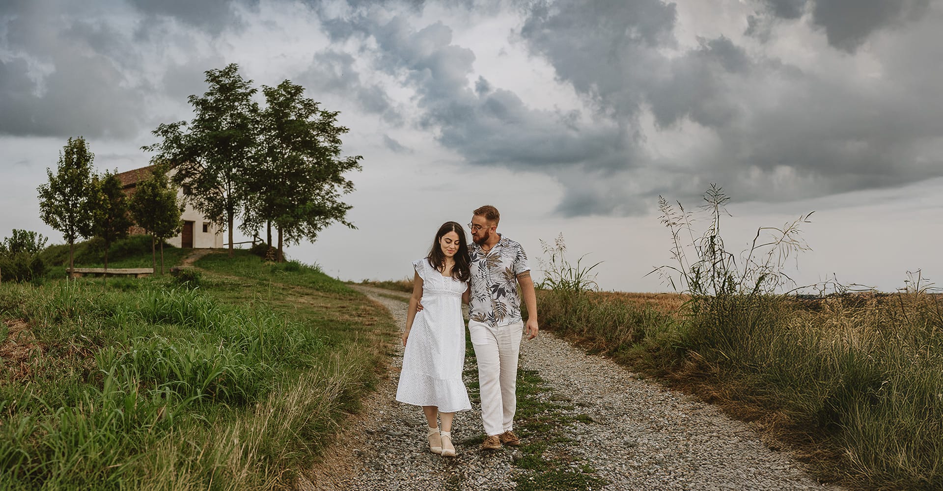 Engagement sulle colline Torinesi. Francesca e Steven camminano abbracciati lungo un sentiero con la chiesetta di San Lorenzo sullo sfondo e il cielo nuvoloso e cupo.