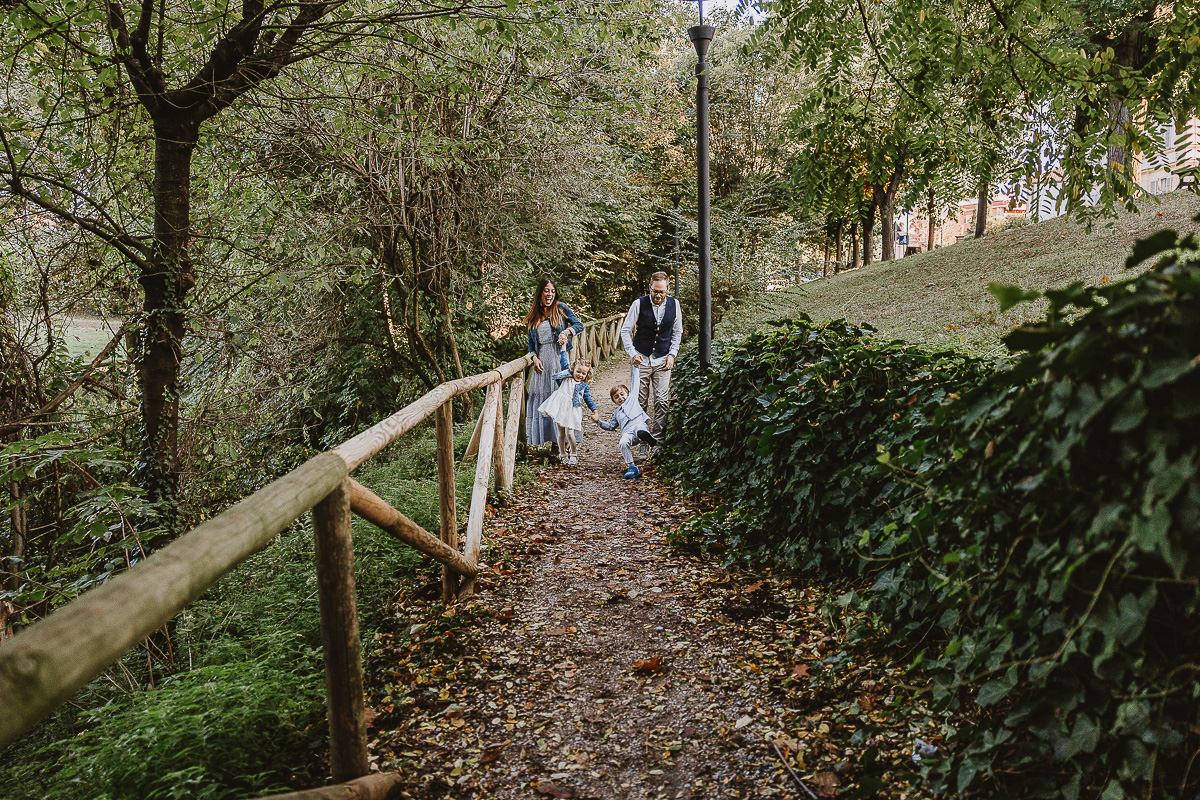 Fotografia di famiglia a Chieri, nel Giardino del Tessile. La famiglia cammina lungo un vialetto e i genitori fanno fare vola vola ai bambini.