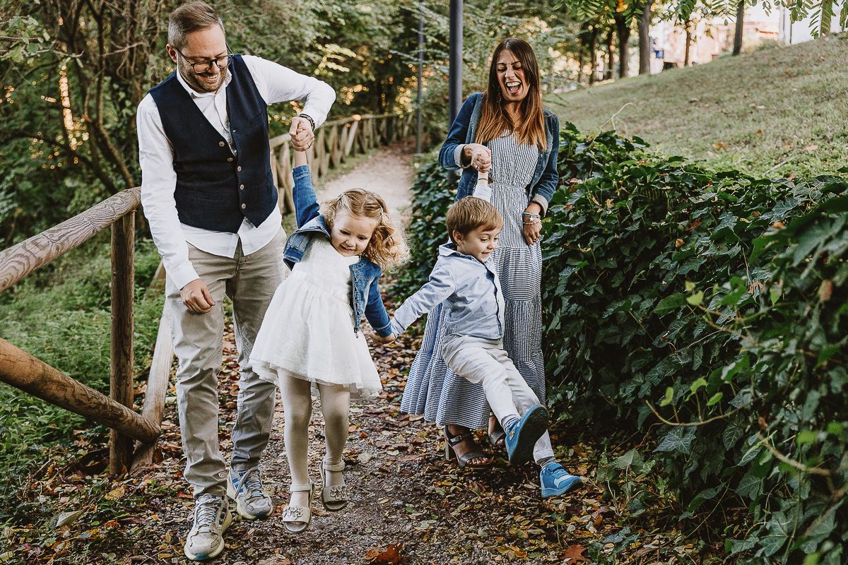 Fotografia di famiglia a Chieri, nel Giardino del Tessile. La famiglia cammina lungo un vialetto e i genitori fanno fare vola vola ai bambini.