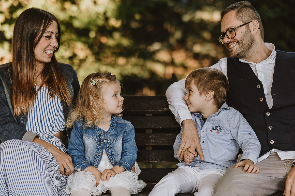 Fotografia di famiglia a Chieri, nel Giardino del Tessile. La famiglia è seduta su di una panchina. I bambini si guardano.
