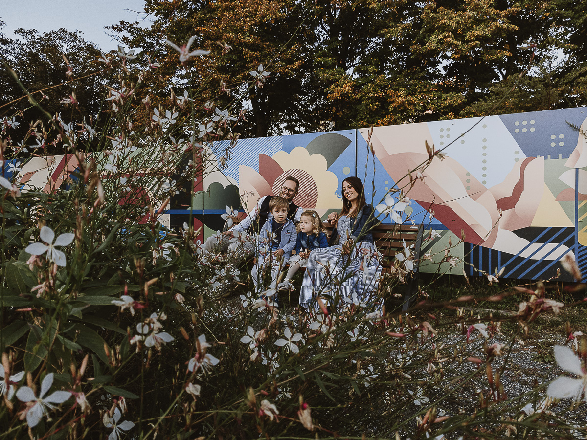 Fotografia di famiglia a Chieri, nel Giardino del Tessile. La famiglia è seduta su di una panchina davanti ai murales. Foto dal drone posto sul terreno.