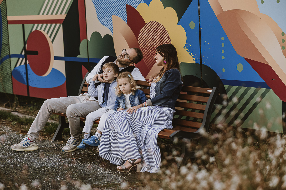 Fotografia di famiglia a Chieri, nel Giardino del Tessile. La famiglia è seduta su di una panchina davanti ai murales.