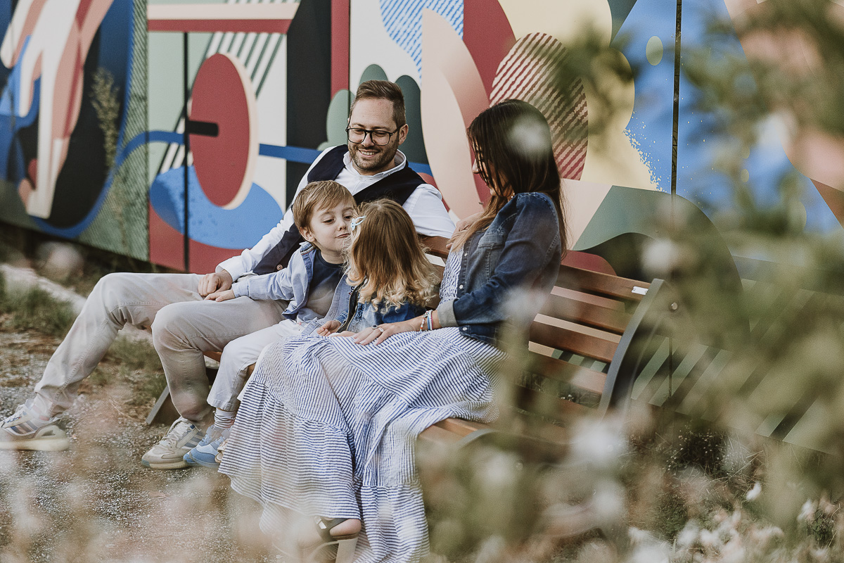 Fotografia di famiglia a Chieri, nel Giardino del Tessile. La famiglia è seduta su di una panchina davanti ai murales.