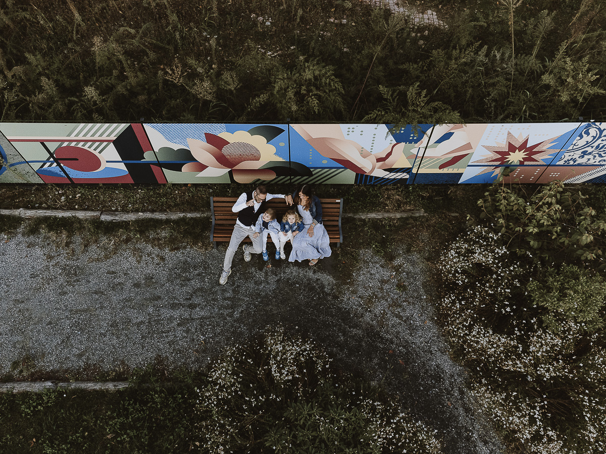 Fotografia di famiglia a Chieri, nel Giardino del Tessile. La famiglia è seduta su di una panchina davanti ai murales. Foto aerea dal drone.