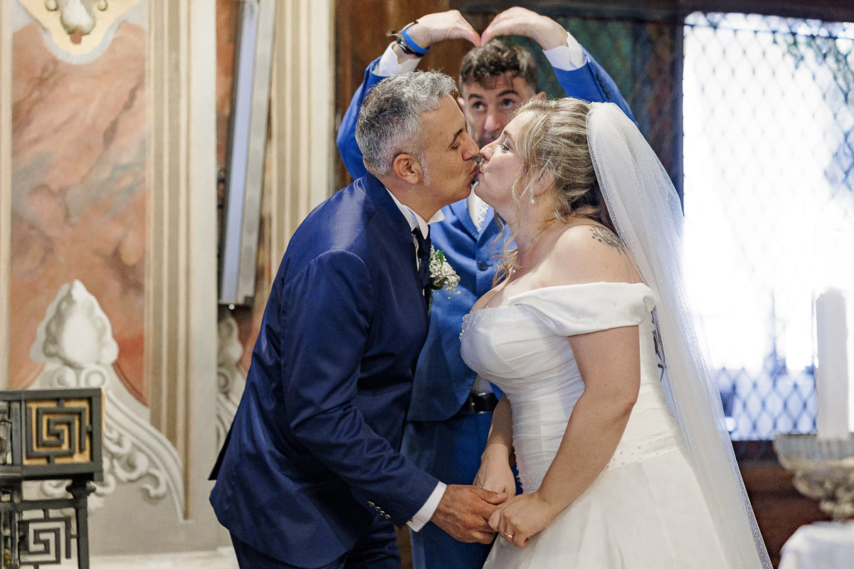 Bacio degli sposi con testimone dietro di loro che fa un cuore con le mani.