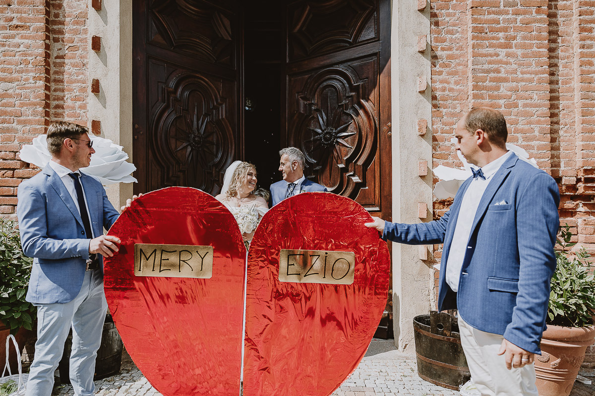 Fotografo Matrimonio in Piemonte. Cerimonia nella chiesa di San Giovanni Battista di Moriondo Torinese. Uscita degli sposi. Gli amici mettono un grande cuore davanti a loro.