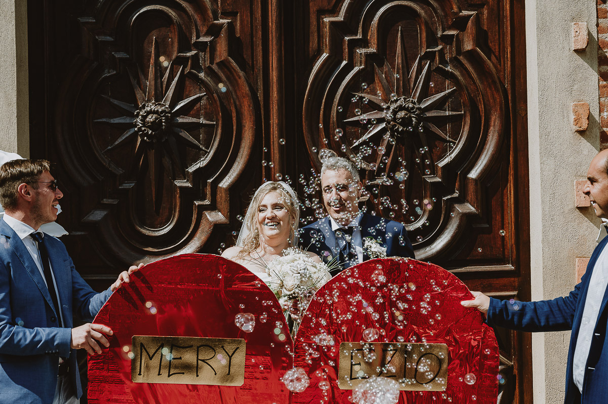 Fotografo Matrimonio in Piemonte. Cerimonia nella chiesa di San Giovanni Battista di Moriondo Torinese. Uscita degli sposi. Gli amici mettono un grande cuore davanti a loro. Lancio delle bolle.