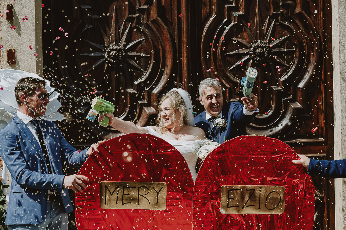Fotografo Matrimonio in Piemonte. Cerimonia nella chiesa di San Giovanni Battista di Moriondo Torinese. Uscita degli sposi. Gli amici mettono un grande cuore davanti a loro. Gli sposi sparano le bolle con due fucili sparabolle.
