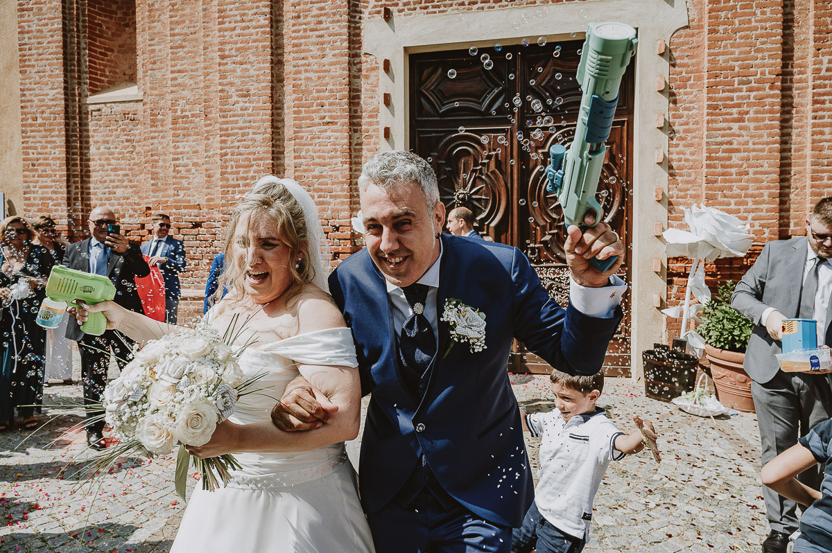 Fotografo Matrimonio in Piemonte. Cerimonia nella chiesa di San Giovanni Battista di Moriondo Torinese. Uscita degli sposi. Gli amici mettono un grande cuore davanti a loro. Gli sposi sparano le bolle con due fucili sparabolle.