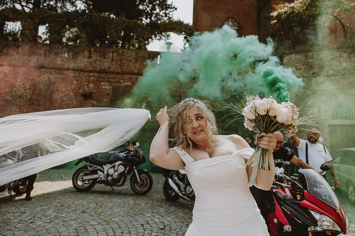 Fotografo Matrimonio in Piemonte. Cerimonia nella chiesa di San Giovanni Battista di Moriondo Torinese. All'uscita gli amici motociclisti hanno creato un cuore con le moto e acceso dei fumi colorati rossi e verdi. Lo sposo tira il velo alla sposa.