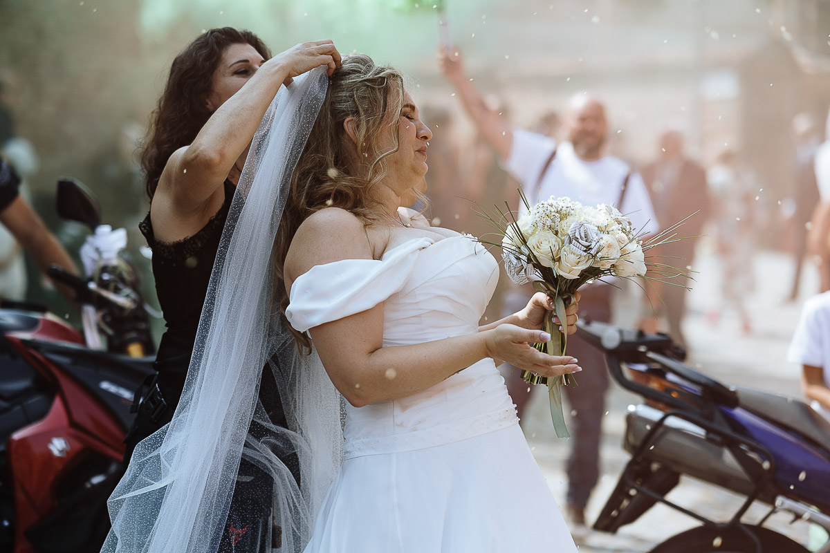 Fotografo Matrimonio in Piemonte. Cerimonia nella chiesa di San Giovanni Battista di Moriondo Torinese. All'uscita gli amici motociclisti hanno creato un cuore con le moto e acceso dei fumi colorati rossi e verdi. Amica rimette il velo alla sposa.