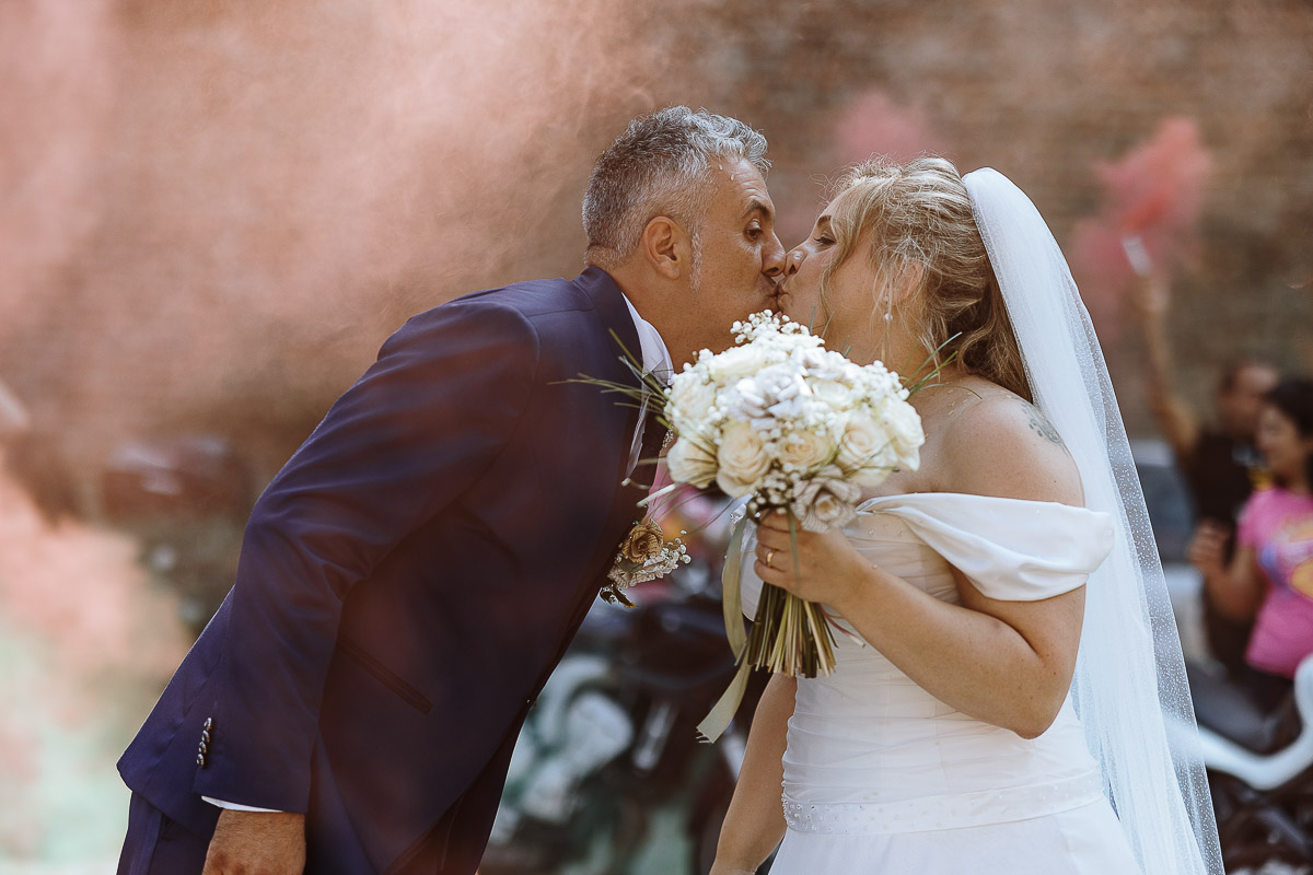 Fotografo Matrimonio in Piemonte. Cerimonia nella chiesa di San Giovanni Battista di Moriondo Torinese. All'uscita gli amici motociclisti hanno creato un cuore con le moto e acceso dei fumi colorati rossi e verdi. Gli sposi si baciano.