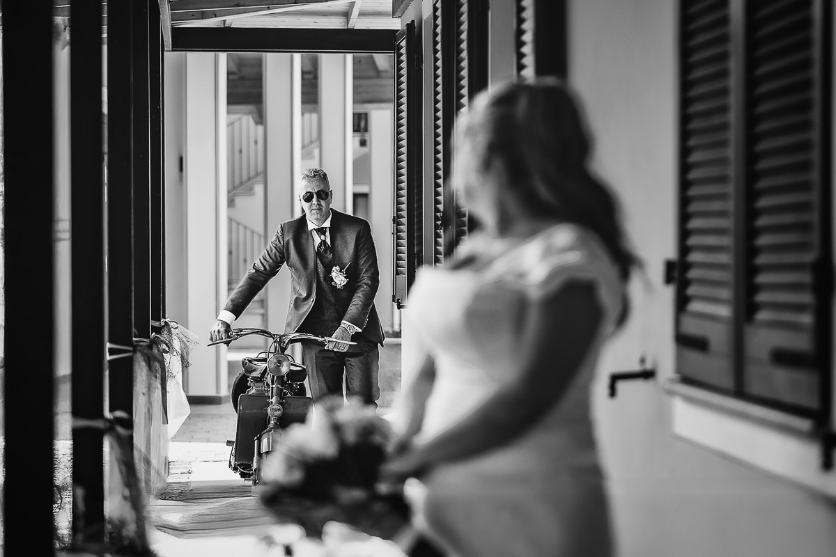 Fotografo Matrimonio in Piemonte. Ricevimento all'agriturismo Greppi di crescentino (VC). Lo sposo arriva con la Lambretta d'epoca. La sposa lo guarda. Bianco e nero.
