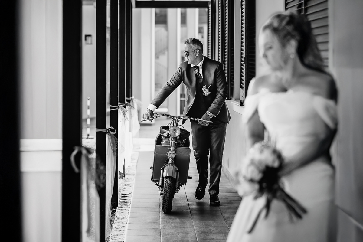 Fotografo Matrimonio in Piemonte. Ricevimento all'agriturismo Greppi di crescentino (VC). Lo sposo arriva con la Lambretta d'epoca. La sposa lo guarda. Bianco e nero.