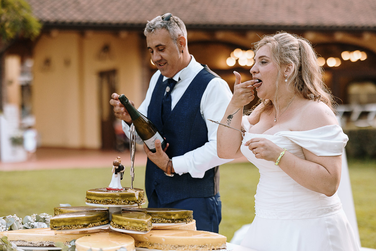 Fotografo Matrimonio in Piemonte. Ricevimento all'agriturismo Greppi di Crescentino (VC). Ricevimento. Taglio torta. Lo sposo apre la bottiglia di spumante e la sposa lecca la paletta taglia torta.