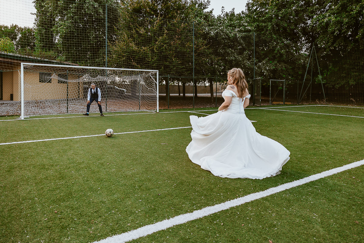 Fotografo Matrimonio in Piemonte. Ricevimento all'agriturismo Greppi di Crescentino (VC). Ricevimento. Ritratti degli sposi al campo di calcio. La sposa calcia il pallone verso la porta difesa dallo sposo.