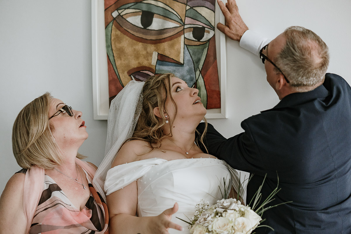 Fotografo Matrimonio in Piemonte. Preparazione a Moncucco Torinese. Sposa con i genitori davanti ad un quadro che sta per cadere.