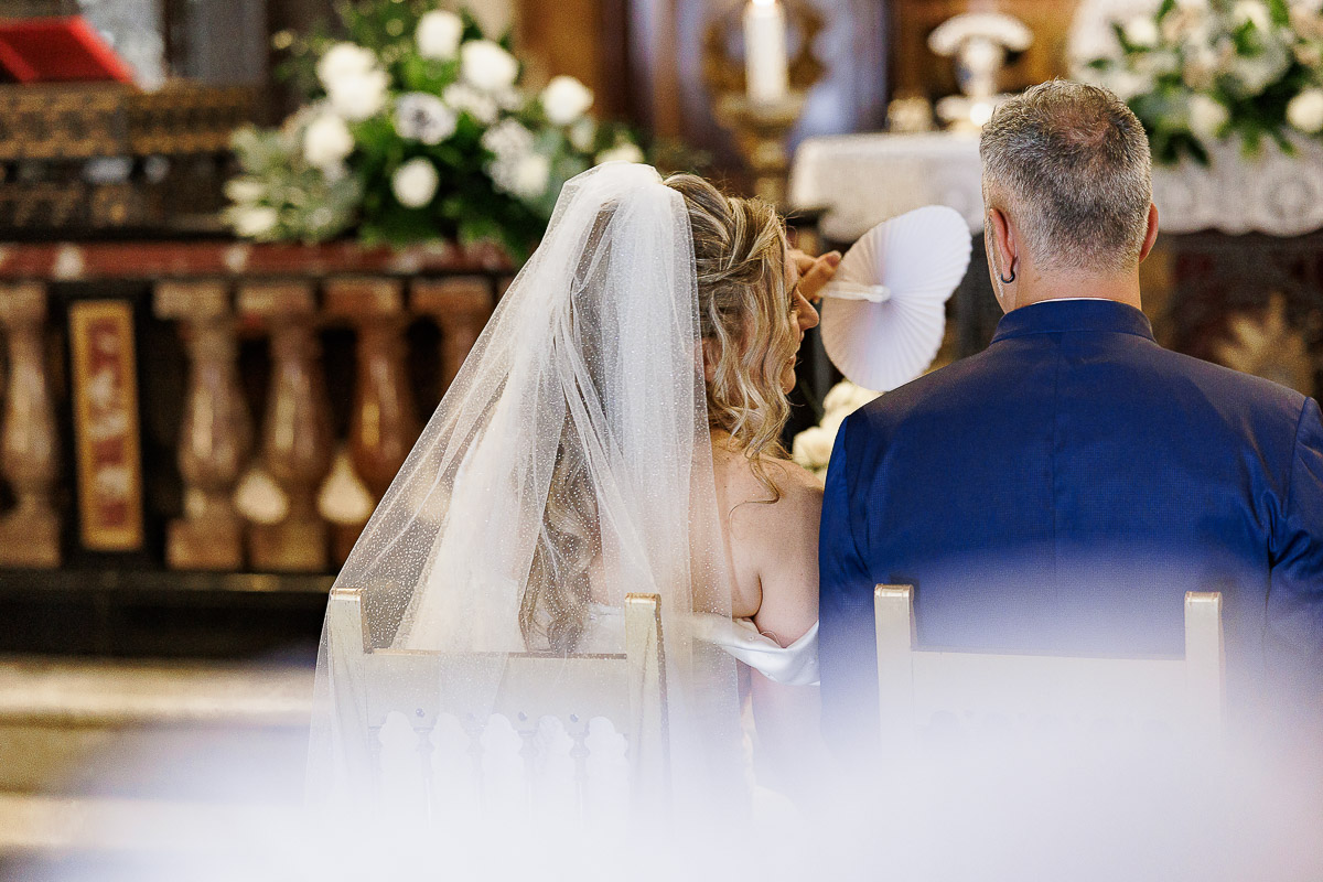 Fotografo Matrimonio in Piemonte. Cerimonia nella chiesa di San Giovanni Battista di Moriondo Torinese. Sposa fa aria allo sposo con il ventaglio a forma di cuore. Visti da dietro.