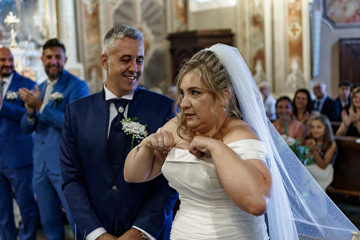 Fotografo Matrimonio in Piemonte. Cerimonia nella chiesa di San Giovanni Battista di Moriondo Torinese. Sposa si sistema il vestito con un'espressione buffa.
