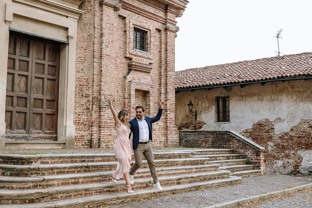 Pre-wedding a Chieri. Shary e Claudio nel piazzale della Chiesa di San Giorgio, il punto più alto e panoramico della città. Shary e Claudio davanti al portone della chiesa scendono le scale. Sltando con le mani alzate.