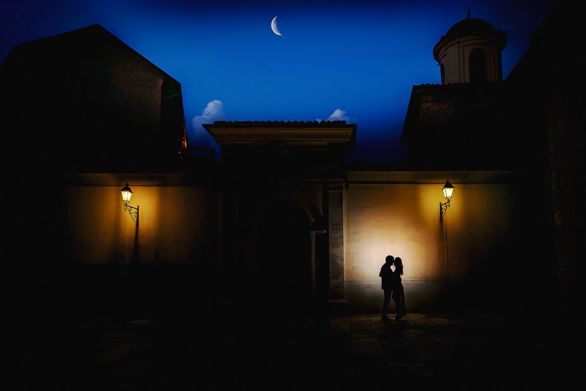 Pre-wedding a Chieri. Shary e Claudio davanti ad un portone, tra due lampioni accesi. Foto serale.. Flash dietro di loro. Silhouette e spicchio di luna nel cielo blu dell'ora blu.