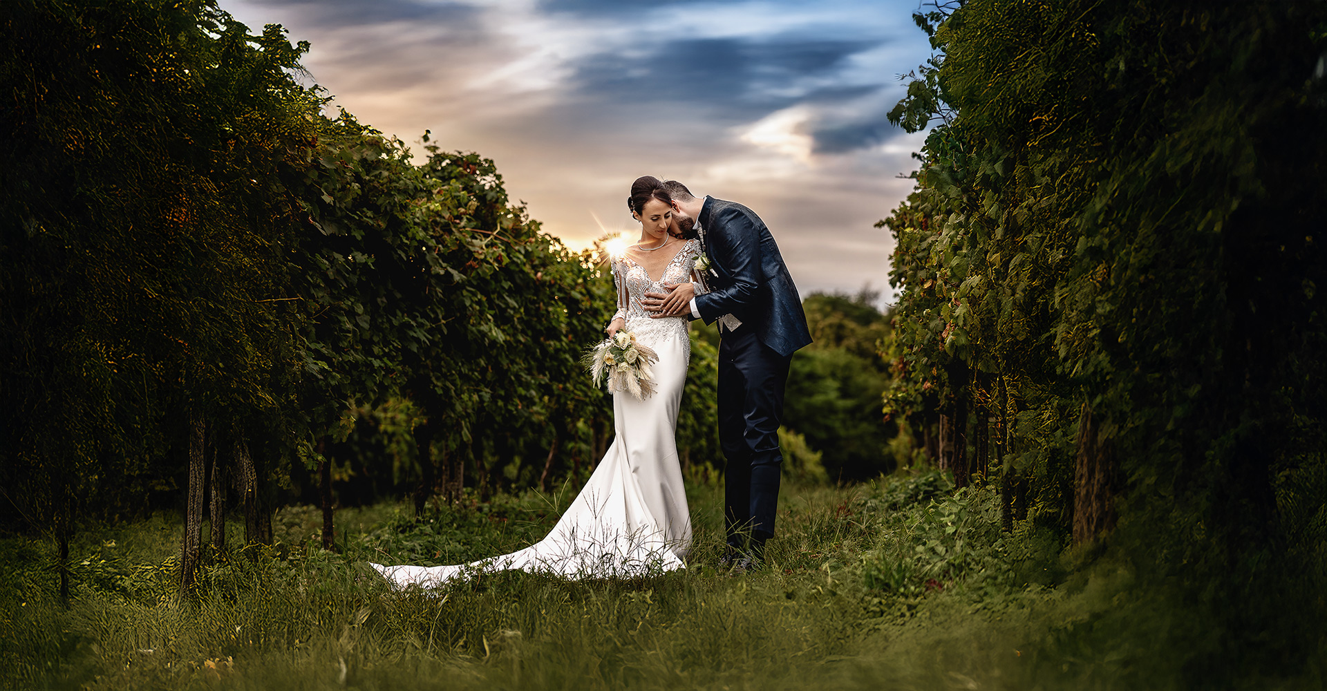 Fotografo matrimonio Lago di Viverone, ritratto degli sposi Ilaria e Luca al tramonto nel Vigneto, Luca bacia sul collo Ilaria mentre un raggio di sole fa capolino dietro la spalla destra della sposa