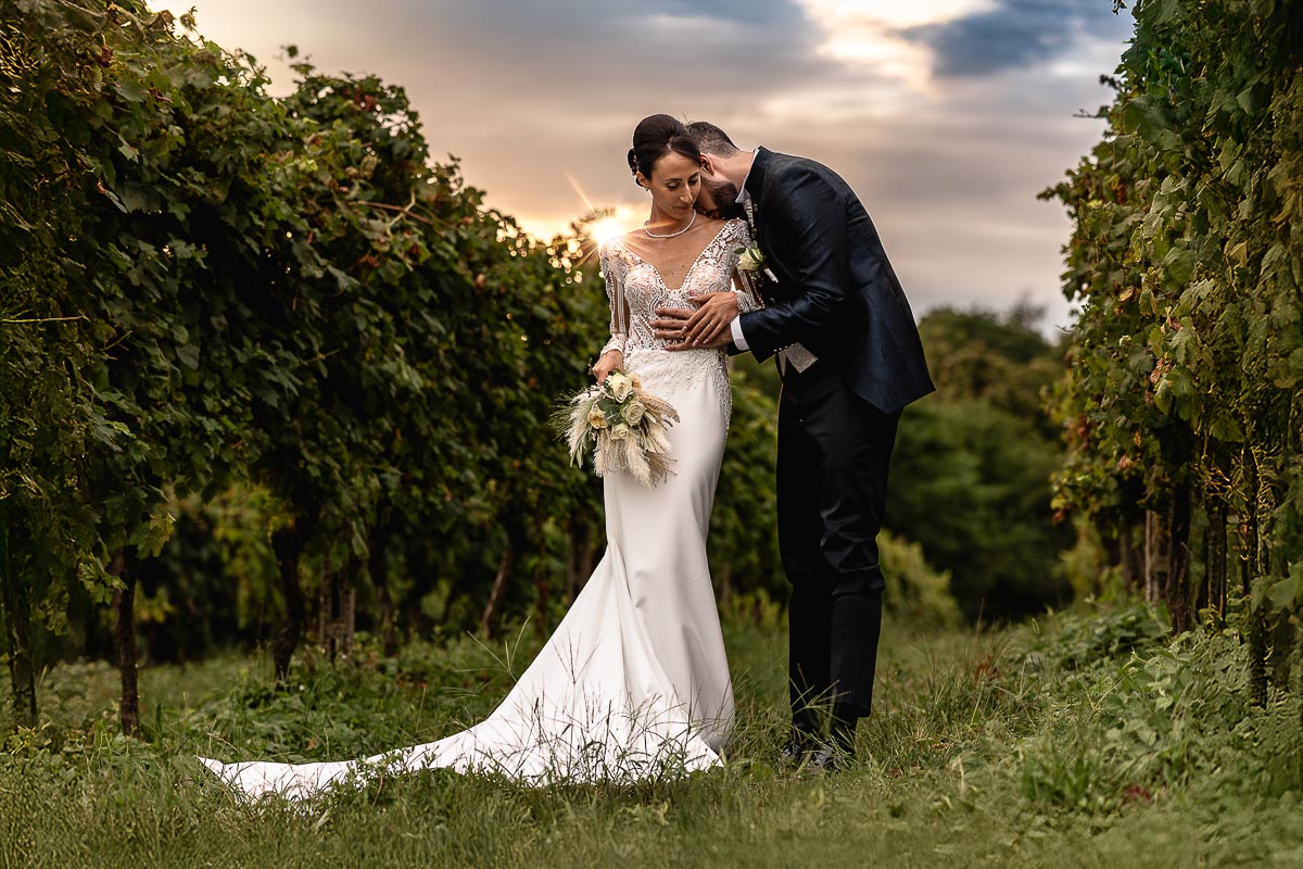 Fotografo Matrimonio Lago di Viverone, Tenuta Variselle. Sessione di coppia preserale nel vigneto. Sposo bacia sposa sulla spalla. Raggio di sole del sole che sta tramontando.