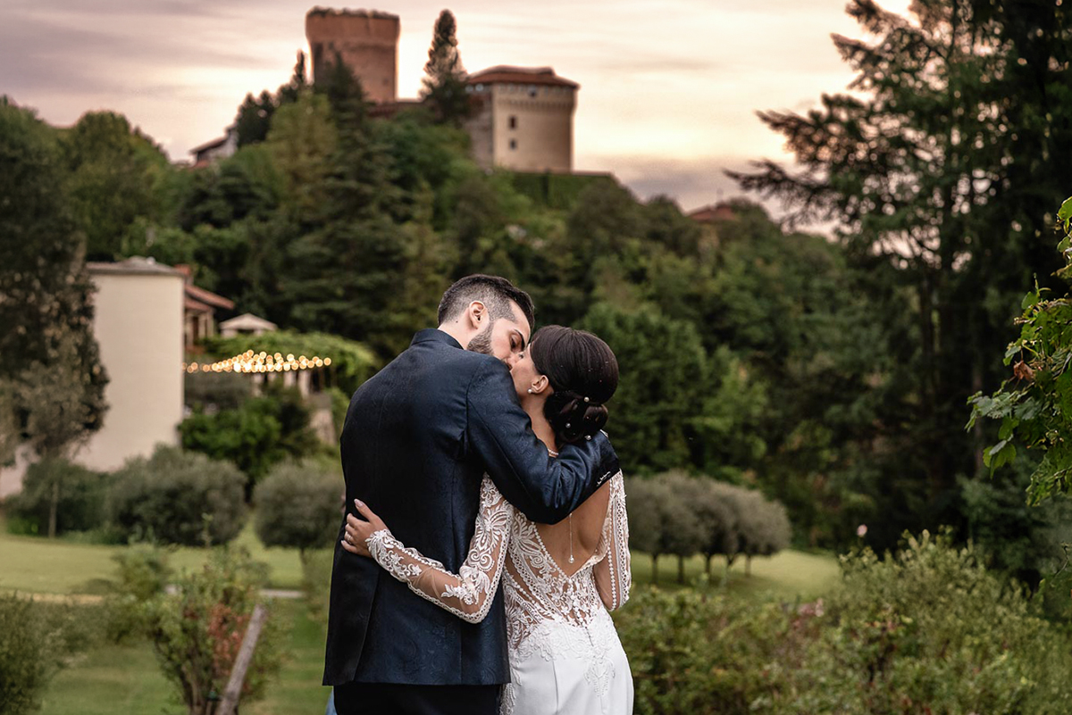 Fotografo Matrimonio Lago di Viverone, Tenuta Variselle. Sessione di coppia preserale nel vigneto. Sposi che si baciano ripresi di schiena. panorama col castello di Roppolo (BI)