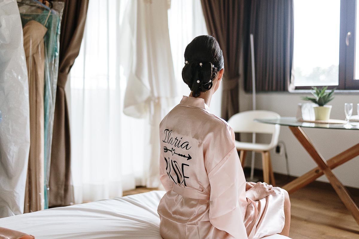 Fotografo Matrimonio Lago di Viverone, Tenuta Variselle. Preparazione della sposa. Sposa seduta sul letto guarda il suo vestito da sposa appeso.