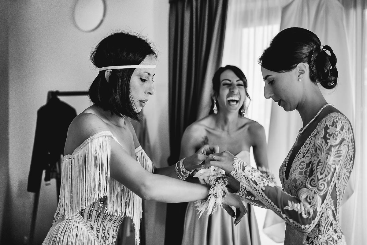 Fotografo Matrimonio Lago di Viverone, Tenuta Variselle. Preparazione della sposa. La sorella aiuta la sposa con le maniche del vestito e l'altra sorella ride.